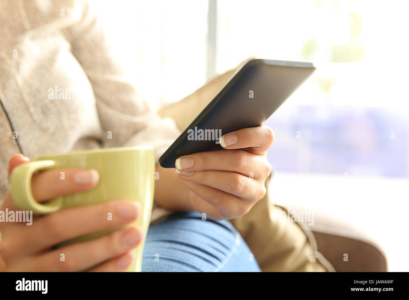 Close up of a relaxed girl hands checking mobile phone on line and holding a cup of coffee sitting on a sofa in the living room at home Stock Photo