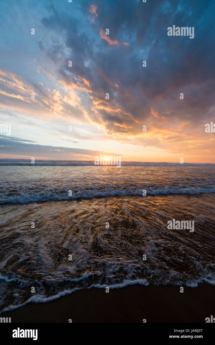 Beneath a storm front an incoming tide pushes waves onto a beach at sunset. Stock Photo