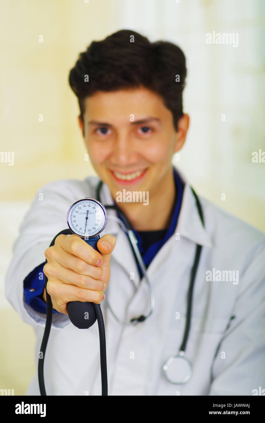 Handsome smiling young doctor with stethoscope around his neck, holdig a Tensiometer in his hand, in a doctor consulting room background Stock Photo