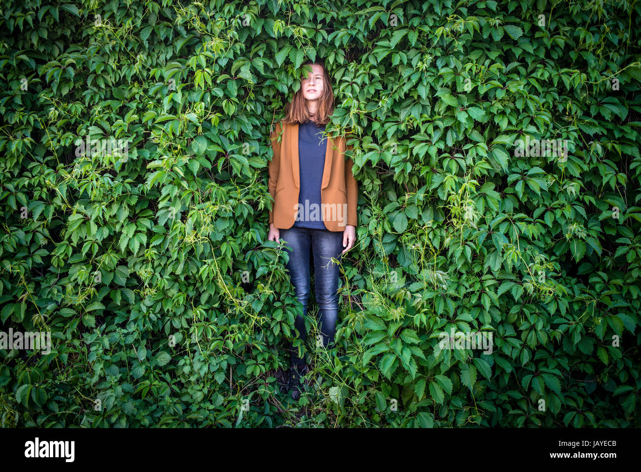A young woman stands near a wall surrounded by greenery Stock Photo