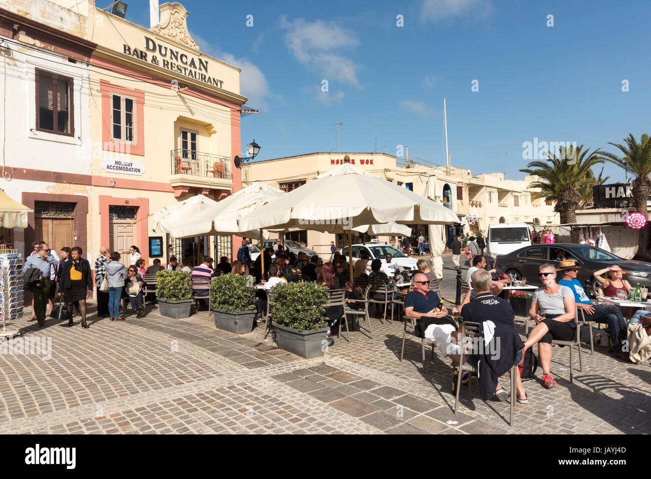 People sitting outside at tables and chairs outside the Duncan bar and restaurant at Marsaxlokk Malta enjoying the sun al fresco Stock Photo