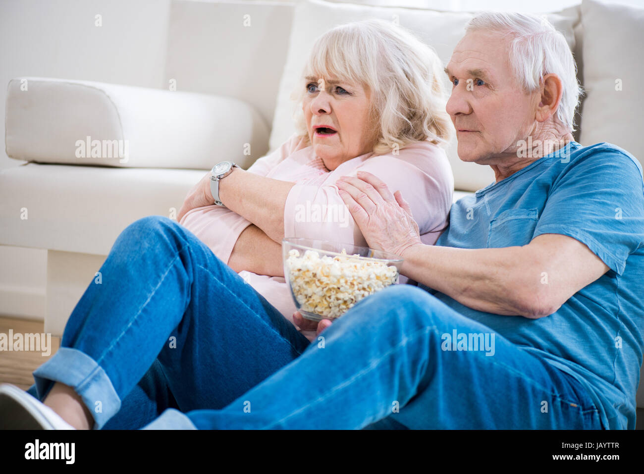 senior couple sitting on floor with popcorn and watching tv, frightened woman. Stock Photo