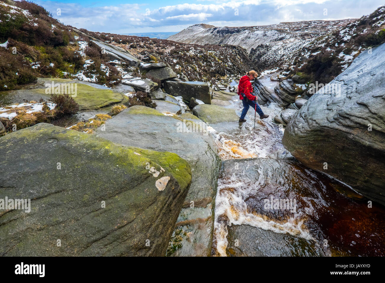 A female walker crossing a stream in winter at the top of Grindsbrook on the southern edge of Kinder Scout, Peak District National Park Stock Photo