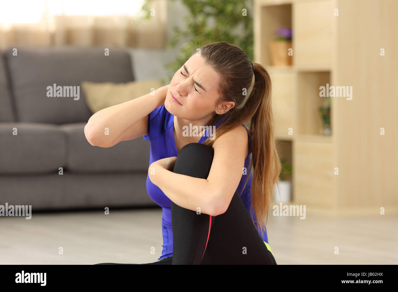 Sportswoman suffering neck ache sitting on the floor in the living room in a house interior with a homey background Stock Photo