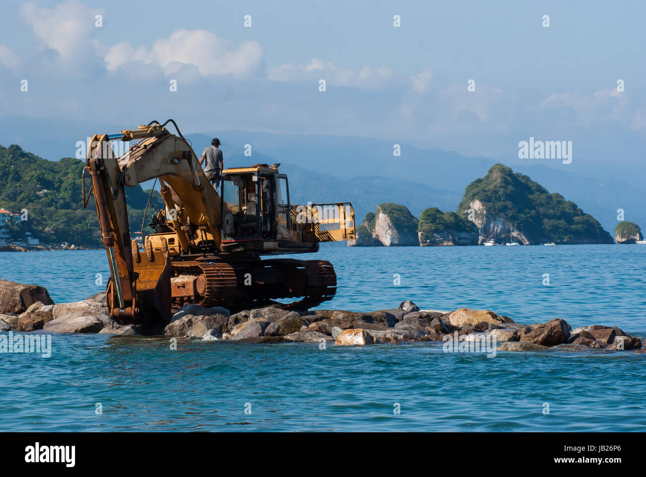 An excavator on a pile of rocks out on the ocean. Los Arcos islands south of Puerto Vallarta in the background. Stock Photo