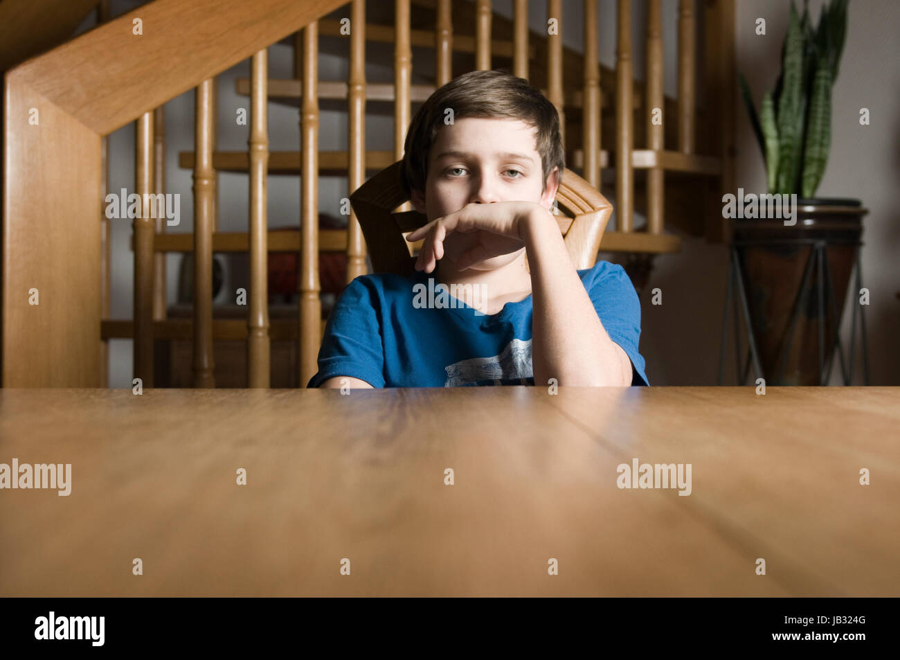 frontale Oberkörper-Ansicht eines jungen männlichen Teenagers im blauen T-Shirt sitzt gelangweilt an einem Holztisch und blickt mit am Mund gehaltener Hand in die Kamera Stock Photo