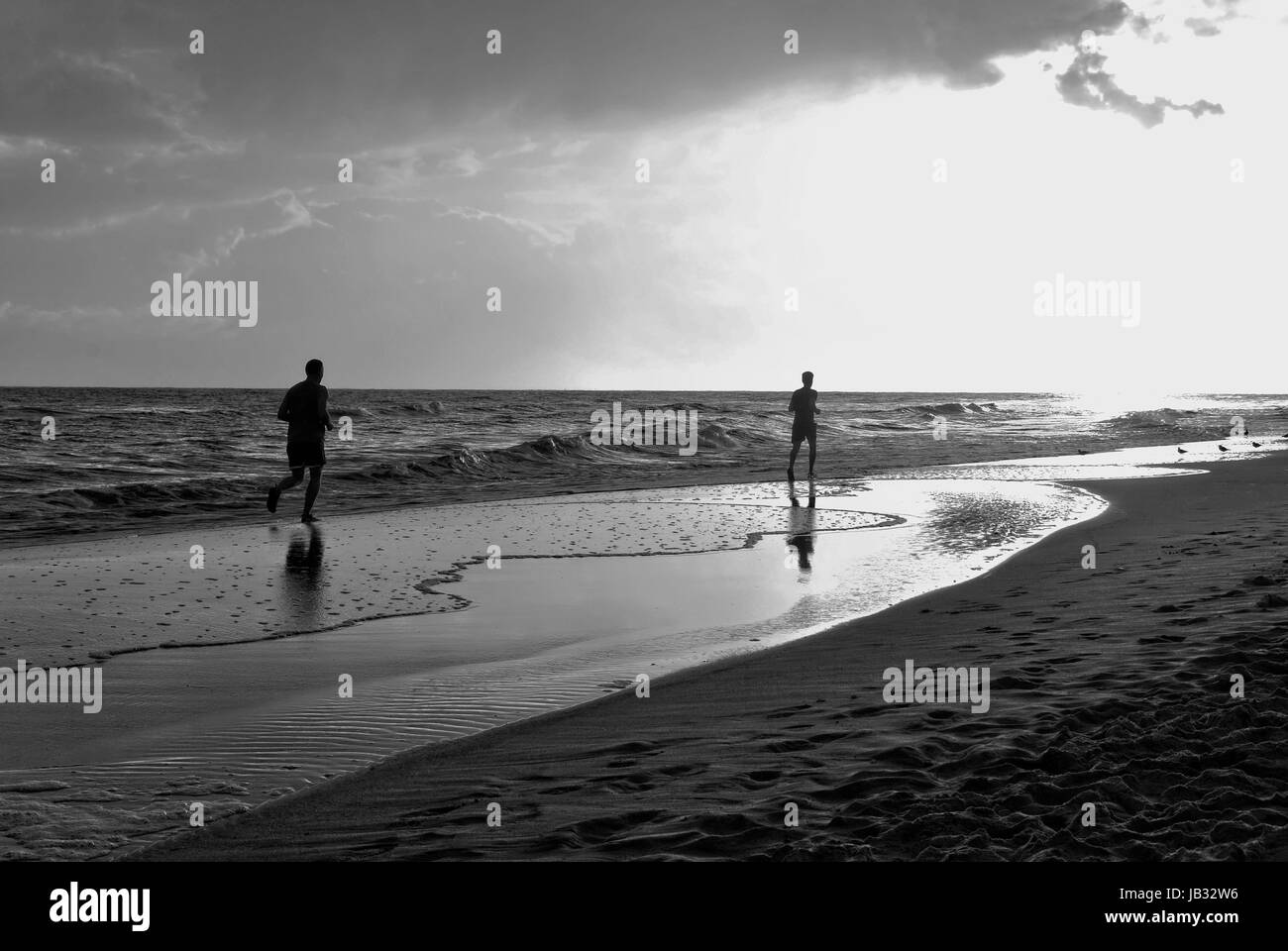 Two men running through the waves towards blinding sun Stock Photo