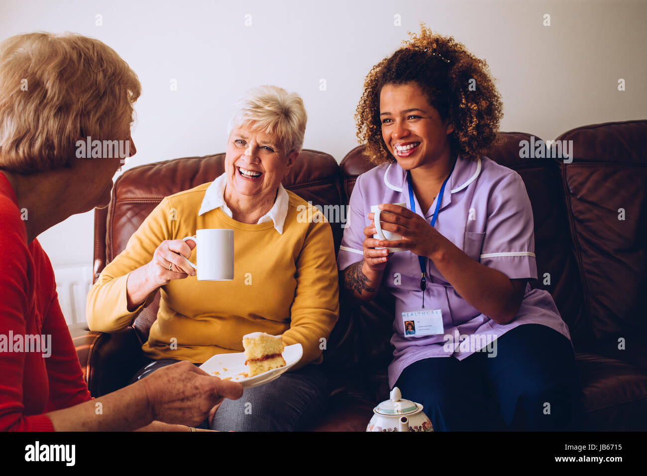 Elderly carer sitting with two of her patients in the care home. They are enjoying some cake and tea. Stock Photo