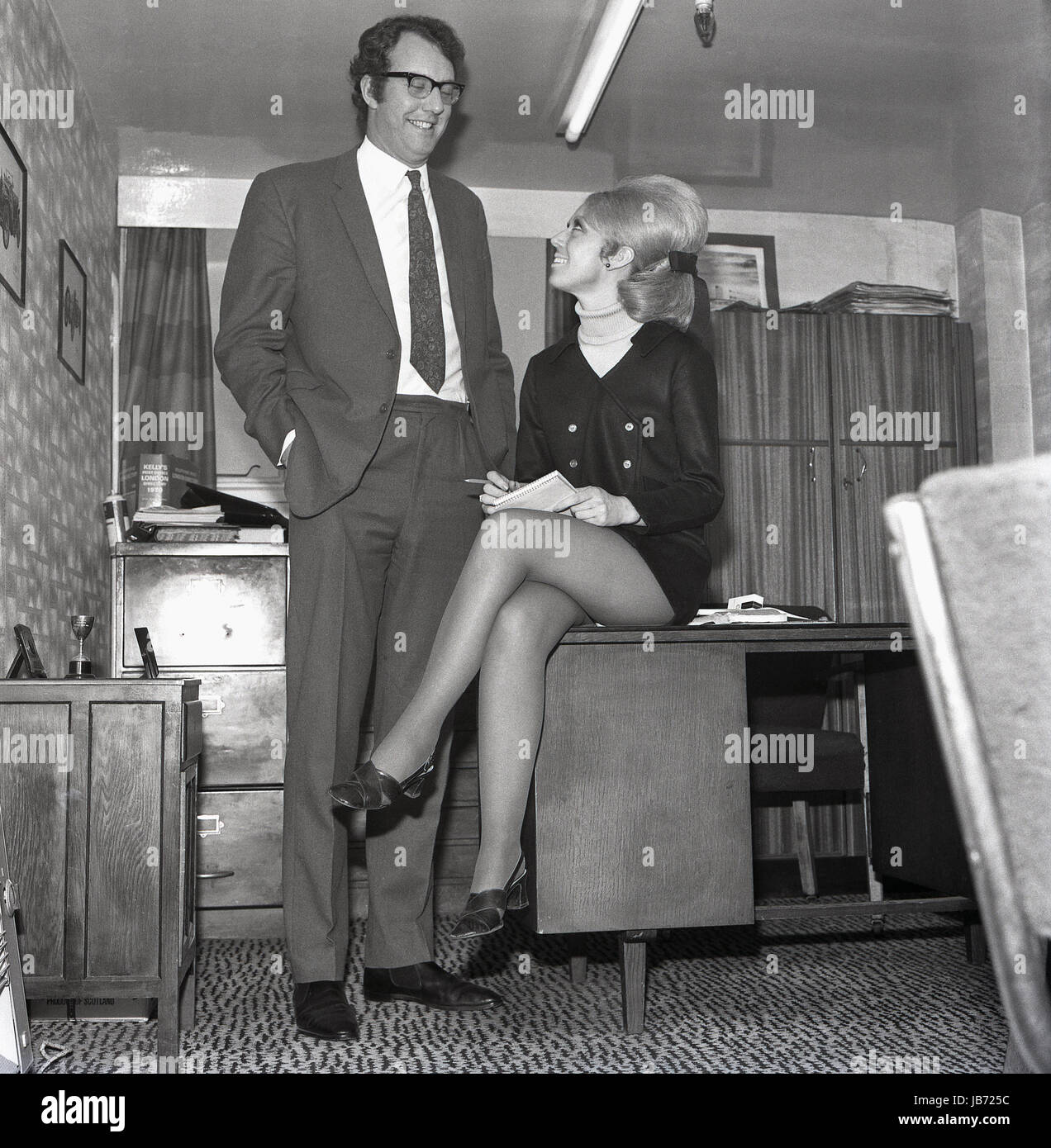 1970s, historical, a secretary wearing a mini-skirt sits on the edge of a  her boss's desk as she takes notes in his office Stock Photo - Alamy