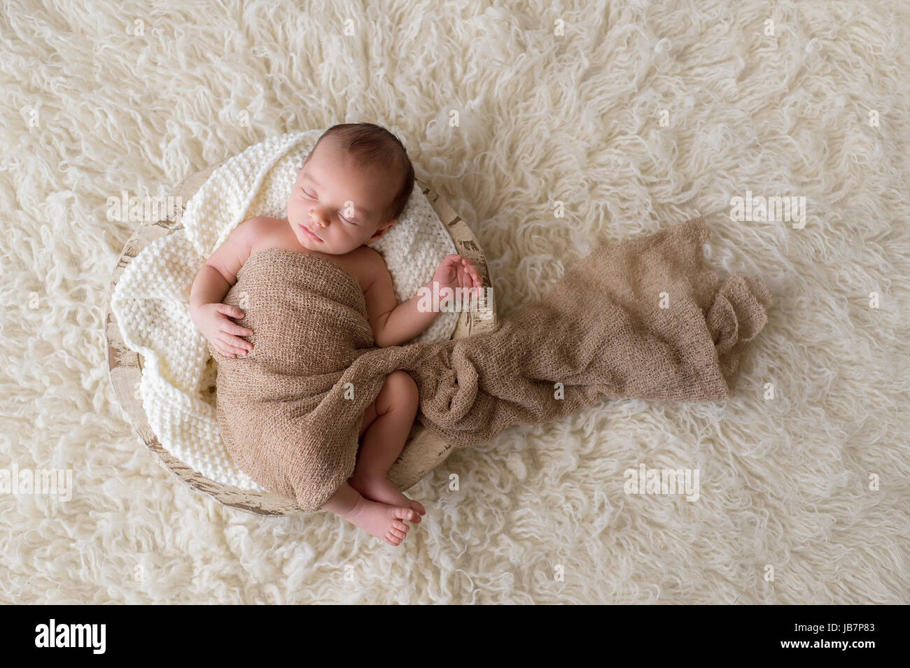 Two week old newborn baby boy swaddled in a beige wrap and sleeping in a round, wooden, trench bowl. Shot in the studio on a cream colored, flokati ru Stock Photo