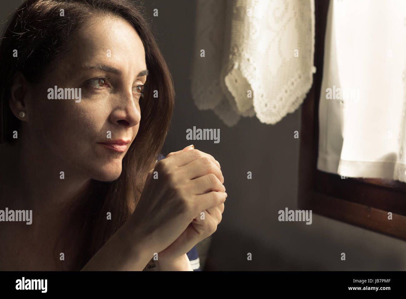 Portrait of a woman looking to a window and hands Intertwined thinking in a positive way, hopeful. Stock Photo