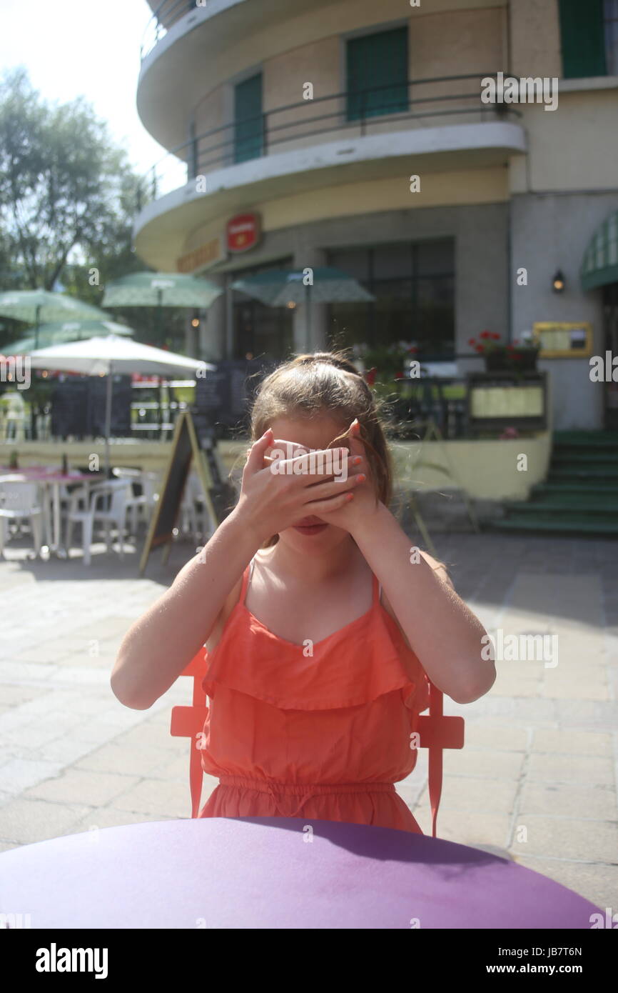 Girl sitting outside a French cafe wearing bright orange summer top with hands crossed over her face - see no evil Stock Photo