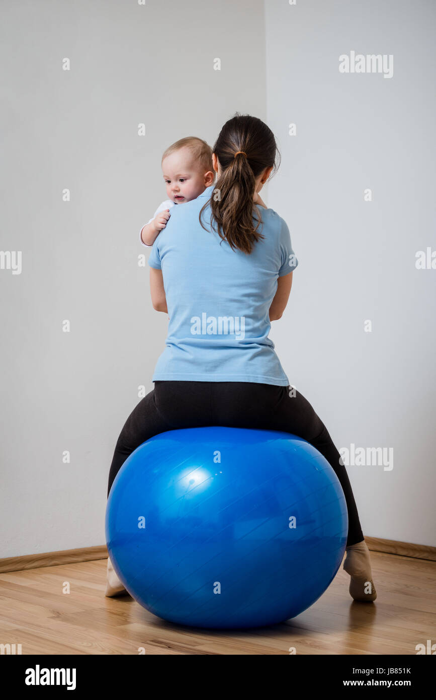Mother exercises with blue fit ball at home holding her baby on hands ...
