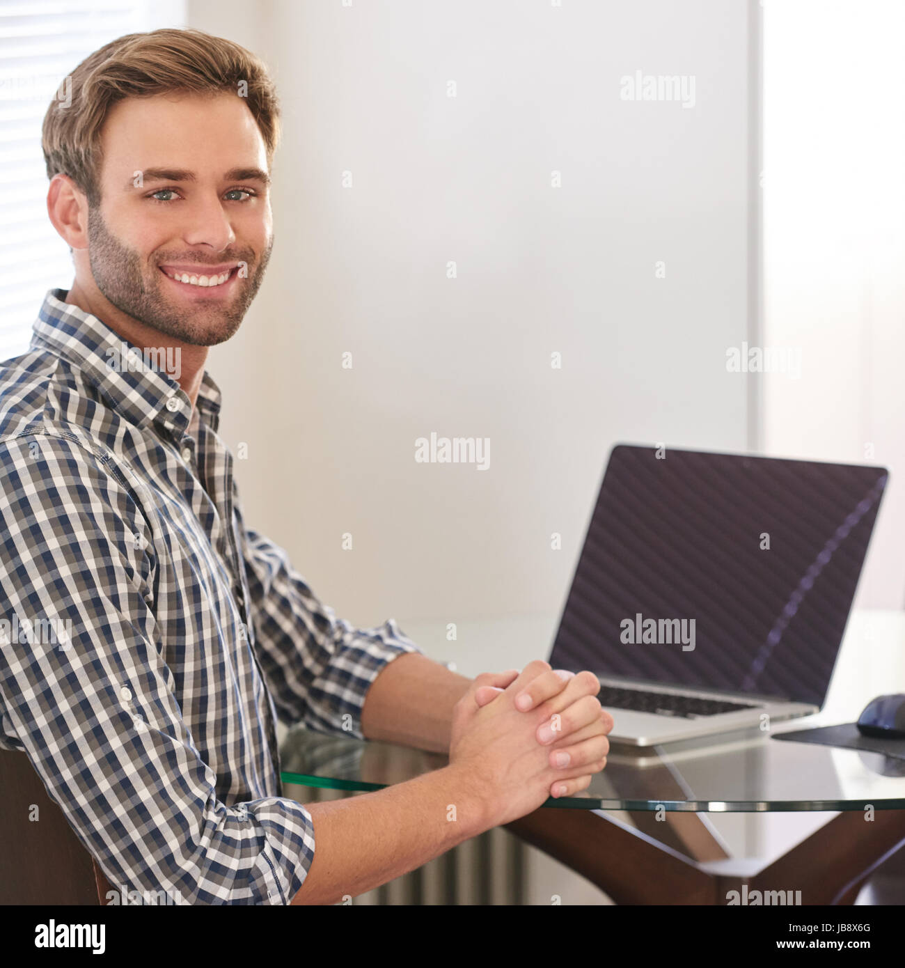 Handsome young man sitting behind his laptop in his living room at a glass table, smiling at the camera over his shoulder with a positive and producti Stock Photo