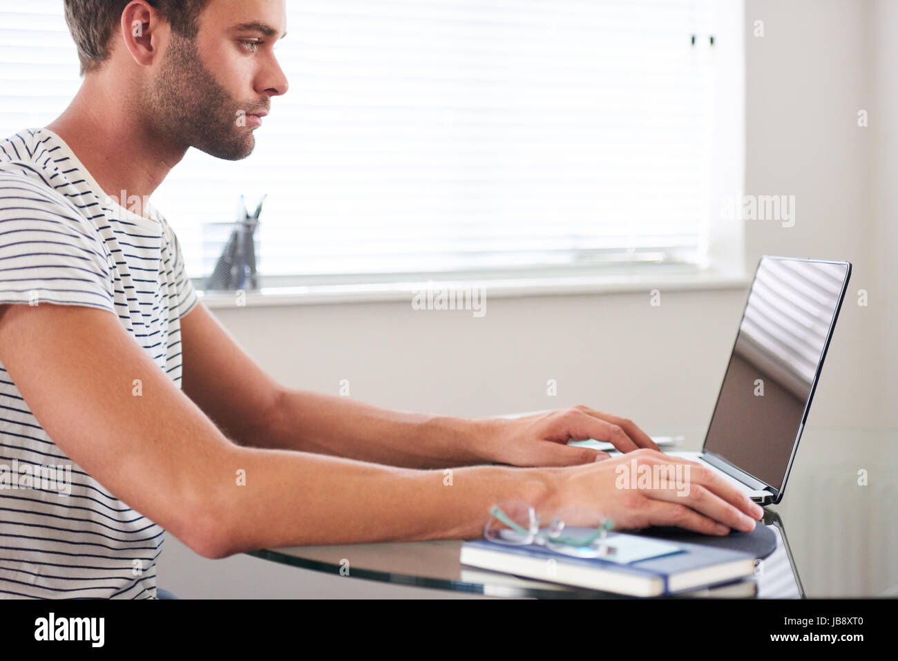 Profile image of young well groomed caucasian male student with a short trimmed beard typing on his computer, seated indoors in his living room next t Stock Photo