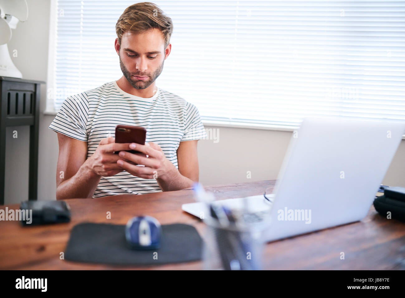 Good looking young caucasian guy busy using his phone with both hands while sitting at home behind his computer at his desk next to a nice large windo Stock Photo