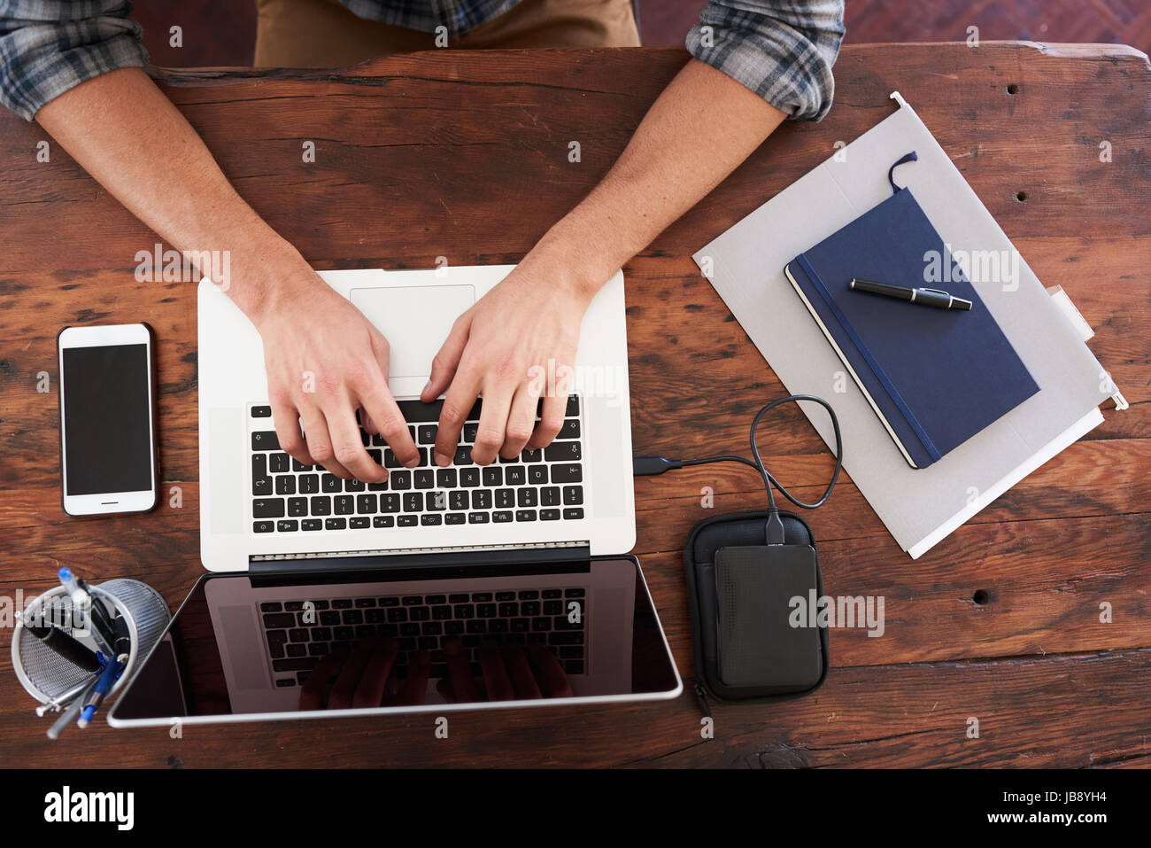 Birds eye view of hands typing on a modern notebook, placed on a durable dark wood desk along with some notes, portable storage, a pen holder and a mo Stock Photo