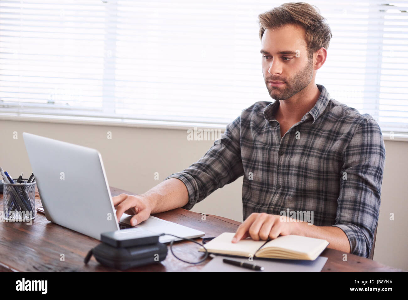 Young caucasian male writer busy digitalising his latest novel from handwritten notebook to a digital format on his modern laptop computer, while keep Stock Photo