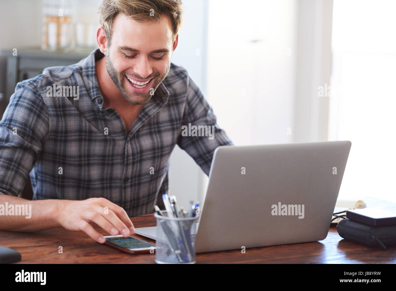 Happy young adult caucasian man smiling joyfully while looking at his phone, reading his latest messages to catch up with his social media as a way of Stock Photo