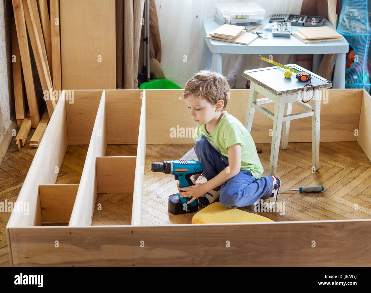 Cute young boy holding screwdriver while sitting on floor at unfinished shelf unit or bookcase Stock Photo