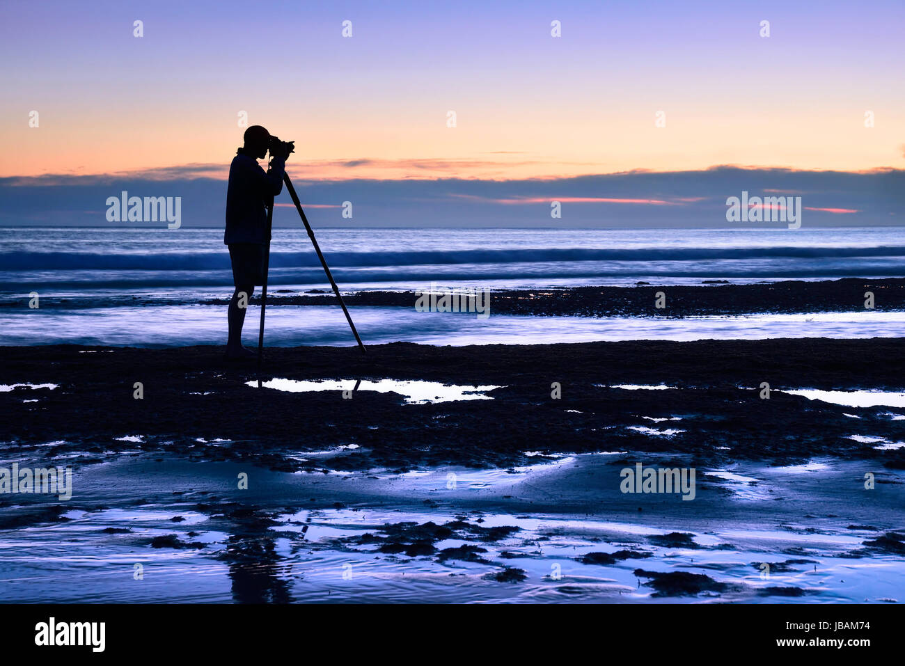 Die Silhouette eines Mannes, der in der Dämmerung am Meer mit Stativ fotografiert Stock Photo