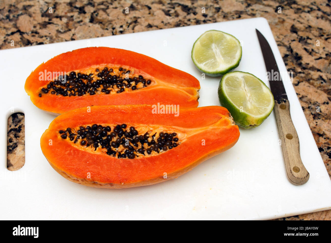 One large dark orange papaya fruit cut in half lengthwise with black seeds  inside next to two halves of a lime on a white cutting board near a knife  on a brown