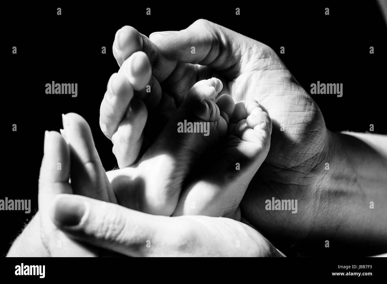 Mammas Haende umfassen zaertlich und beschuetzend Babys kleine Fuesse, freigestellt vor dunklem Hintergrund. Stock Photo