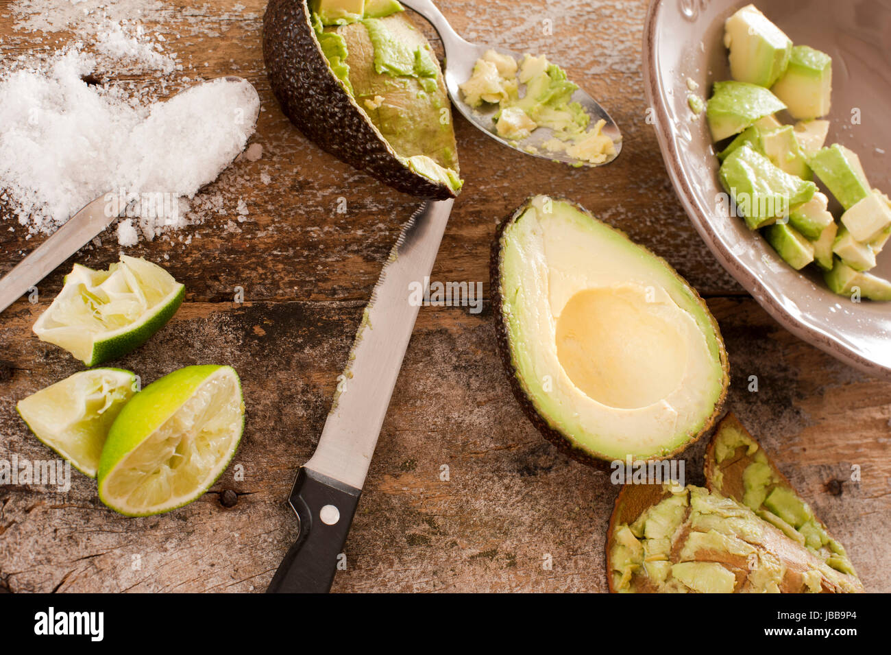 Preparing a delicious fresh avocado salad with a halved avocado next to a bowl with peeled and diced pulp together with a knife, salt and squeezed lemon on an old wooden table, view from above Stock Photo