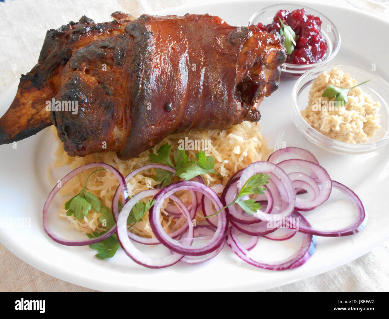 Traditional Czech dish of roasted pork leg with fresh red onions, horseredish, sauerkraut, cranberry sauce on a white plate. Stock Photo