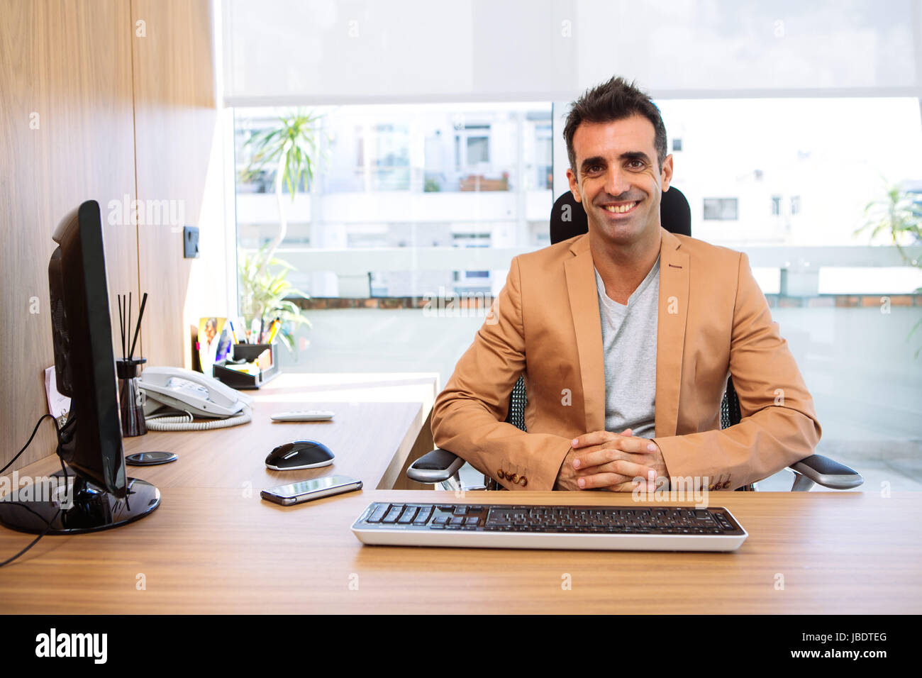 A 35 - 40 years man caucasian dark hair cool modern informal look smiling seating behind a desk in a work station in an office Stock Photo
