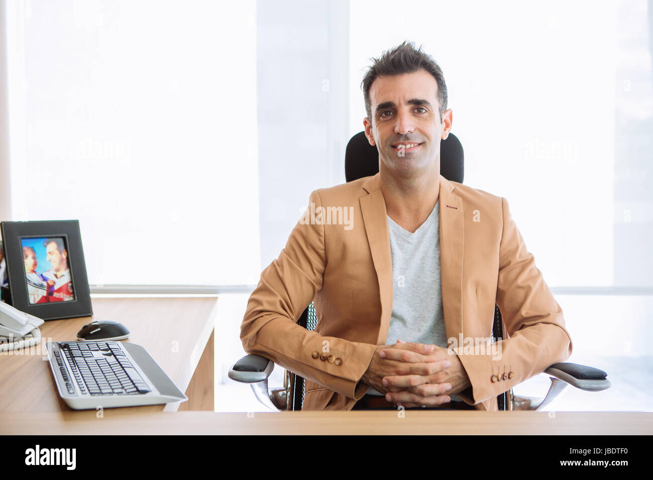 A 35 - 40 years man caucasian dark hair cool modern informal look smiling seating behind a desk in a work station in an office Stock Photo