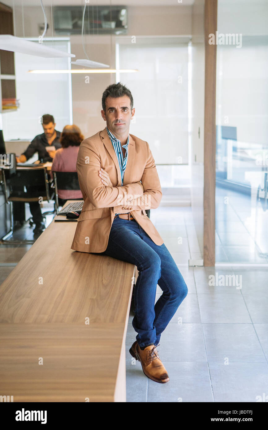 A 35 40 years old man caucasian dark hair cool modern look arms folded smiling in a stripped shirt blazer and jeans seating on a desk in an office Stock Photo Alamy