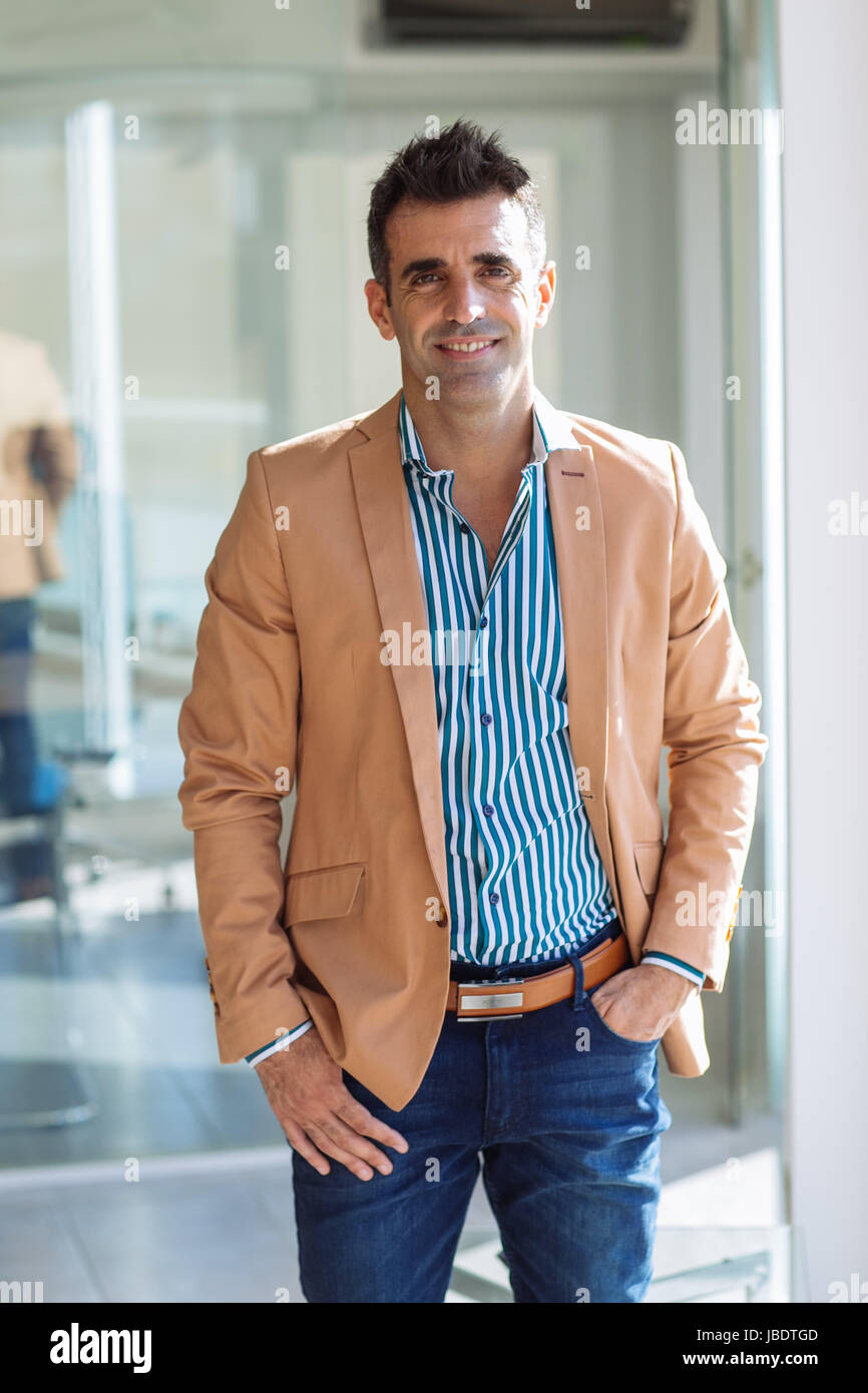 A 35 - 40 years old man caucasian dark hair cool modern look smiling in a stripped shirt, blazer and jeans in an office, medium long shot Stock Photo