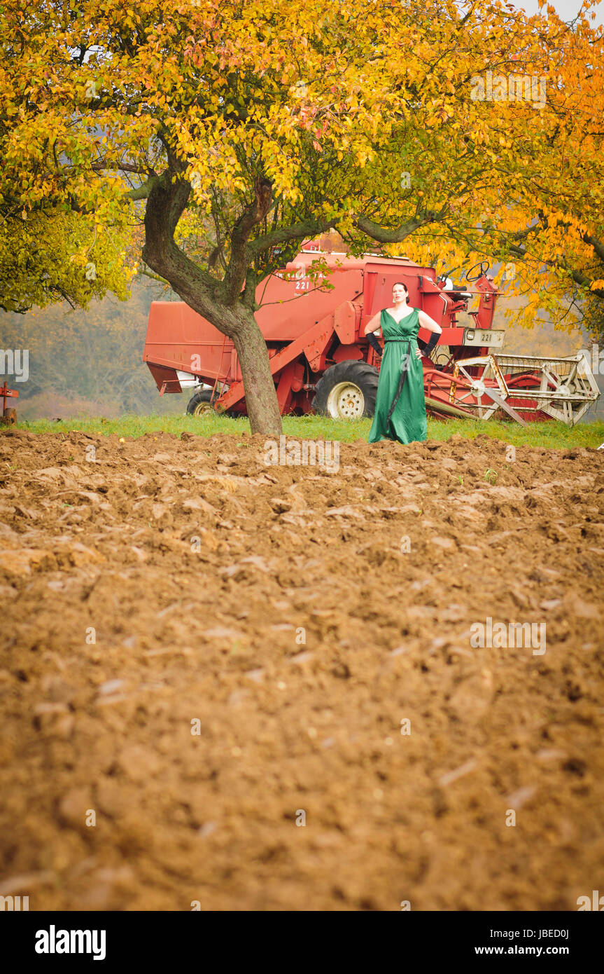 Erwachsene Frau im eleganten Abendkleid arbeitet in der Landwirtschaft und schaut erwartungsvoll auf ein frisch gepfügtes Feld. Stock Photo