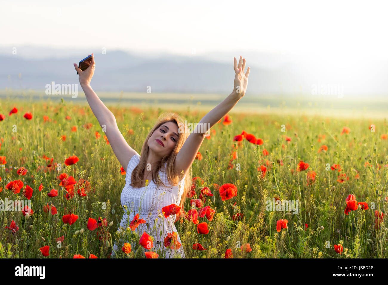 Young girl wearing white dress cloth standing in a poppy field with hands up with phone Stock Photo