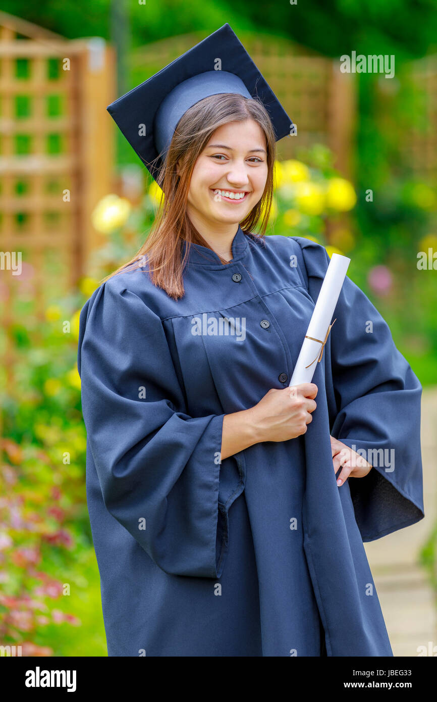 Smiling young woman holding diploma and wearing cap and gown outdoors looking at camera. Graduation concept. Stock Photo
