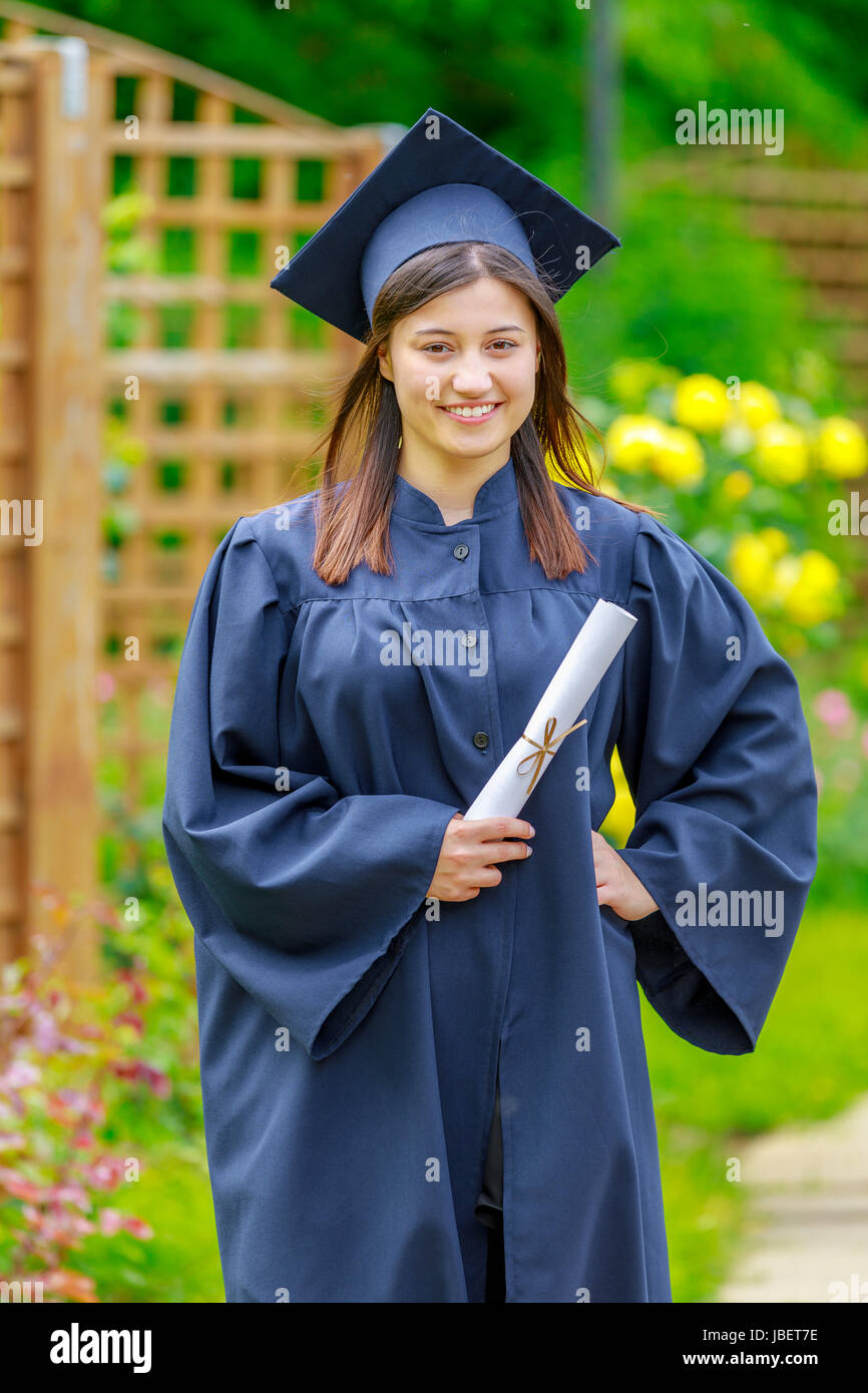 Smiling young woman holding diploma and wearing cap and gown outdoors looking at camera. Graduation concept. Stock Photo