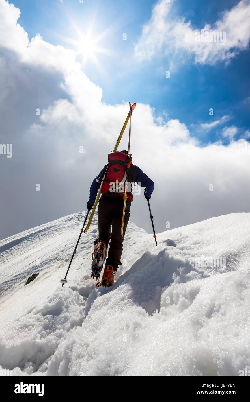Ski mountaineer walking up along a steep snowy ridge with the skis in the backpack. In background a dramatic sky with a shiny bright sun. Concepts: adventure, achievement, courage, determination, self-realization, dangerous activity, extreme sport, winter leisure. Stock Photo