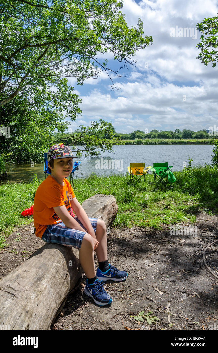 A young boy sits on a log at a campsite next o The River Thames in Berkshire. Stock Photo