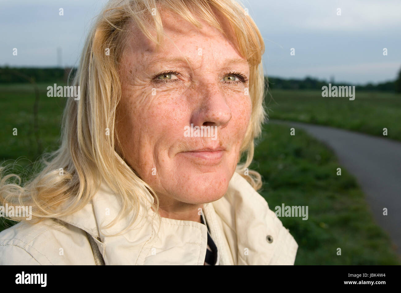Kopf-und-Schulter-Porträt einer blonden reifen Frau in heller Jacke auf einer grünen Wiese mit Fußweg in die Kamera blickend Stock Photo