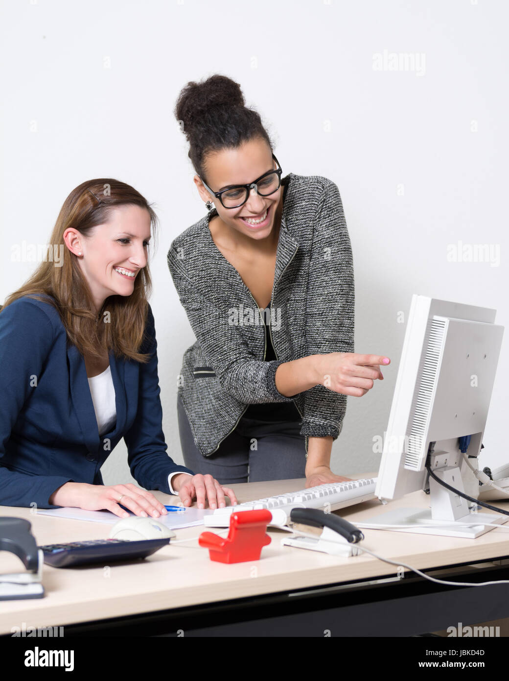 Zwei Frauen stehen bzw. sitzen vor dem Computer im Büro. Beide Frauen schauen zum Monitor und lächeln. Eine Frau zeigt zum Monitor. Stock Photo