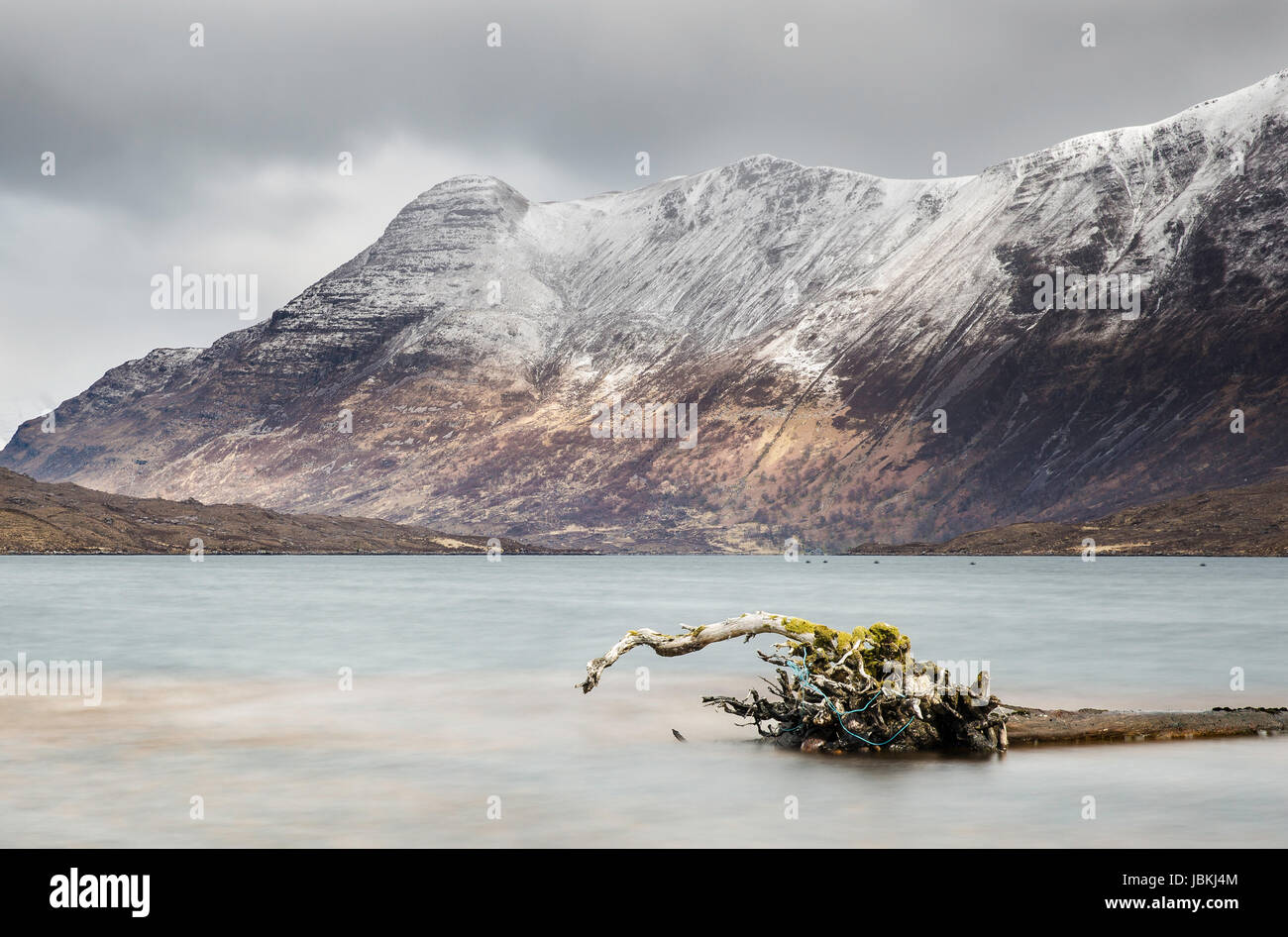 Loch Damh in Torridon, Scotland, with snowy mountains in the distance and an old tree truck floating in the lake in the foreground Stock Photo