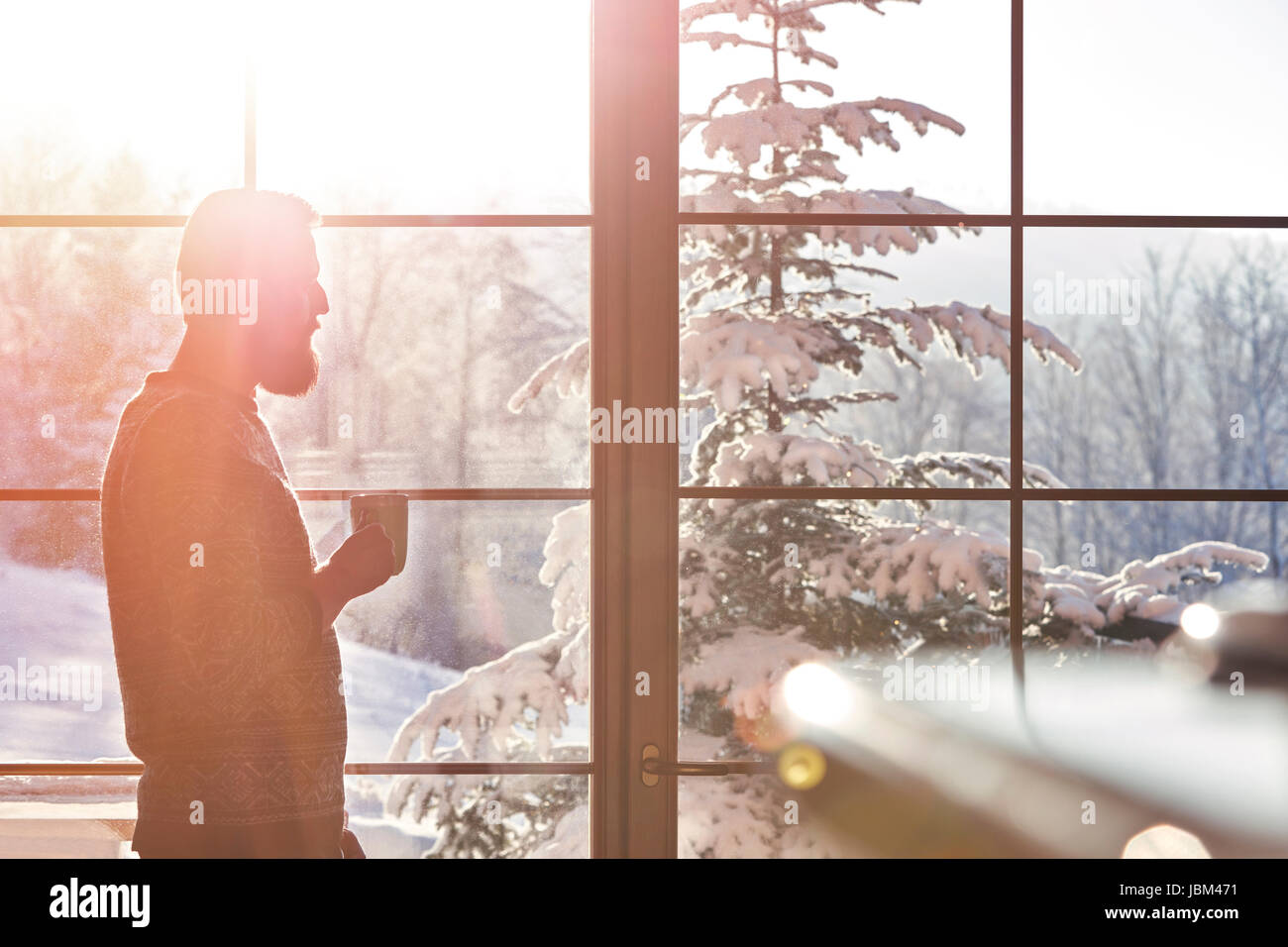 Man drinking coffee at sunny window with view of snowy trees Stock Photo