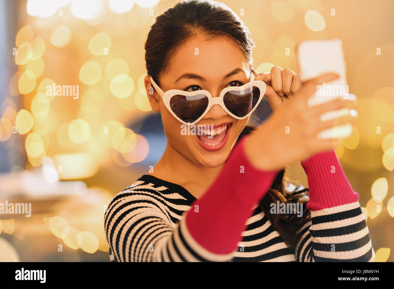 Playful Chinese woman wearing heart-shape glasses taking selfie with camera phone Stock Photo