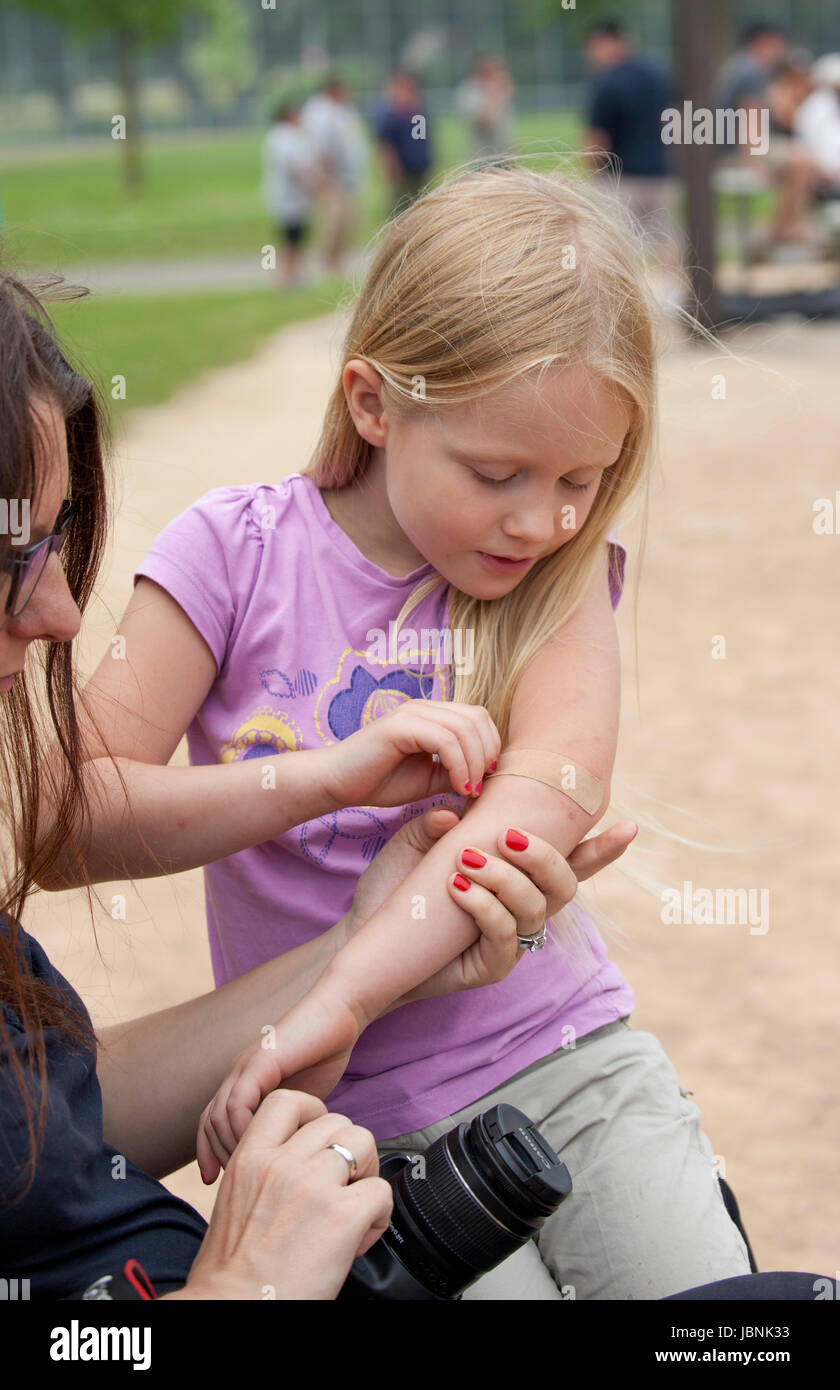 Mom helps daughter age 7 cover bug bites on her arm with a bandage at city park. St Paul Minnesota MN USA Stock Photo