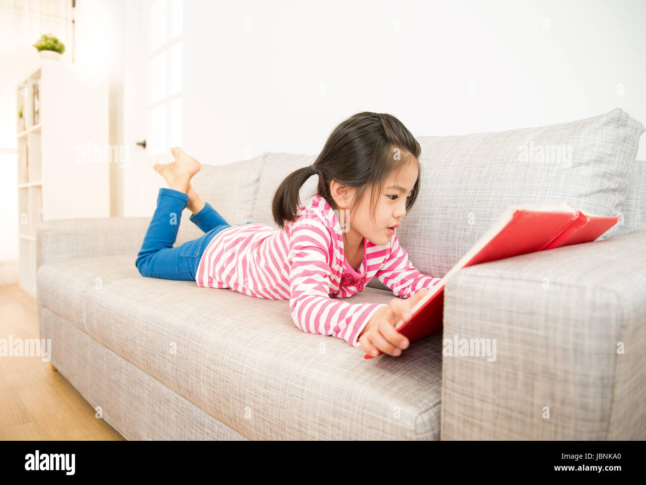 happy little girl lying down on sofa reading story book in the living ...