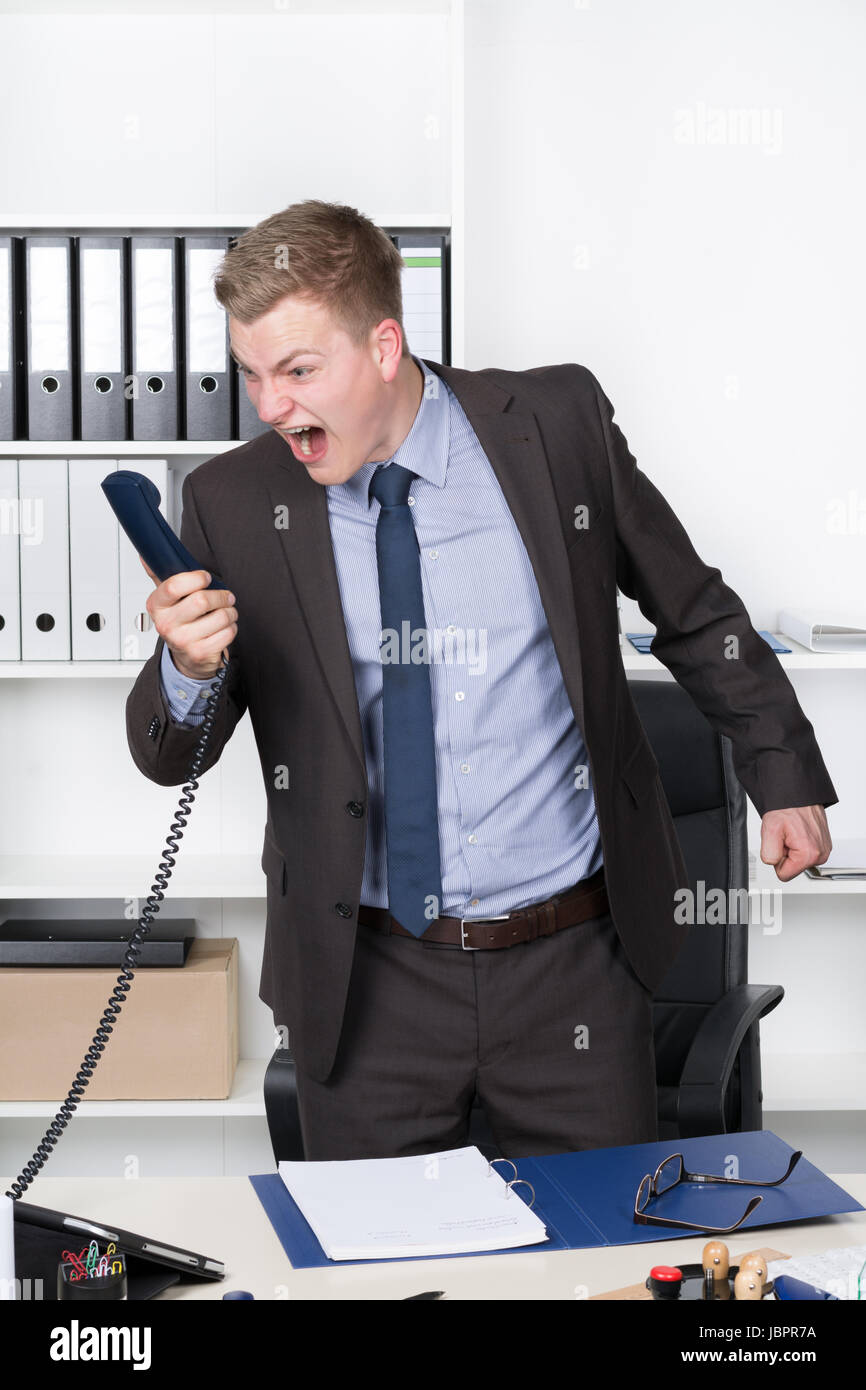 Junger Geschäftsmann steht am Schreibtisch im Büro und schreit ins Telefon. Im Hintergrund befindet sich ein Regal. Der Mann schaut zum Telefonhörer. Stock Photo