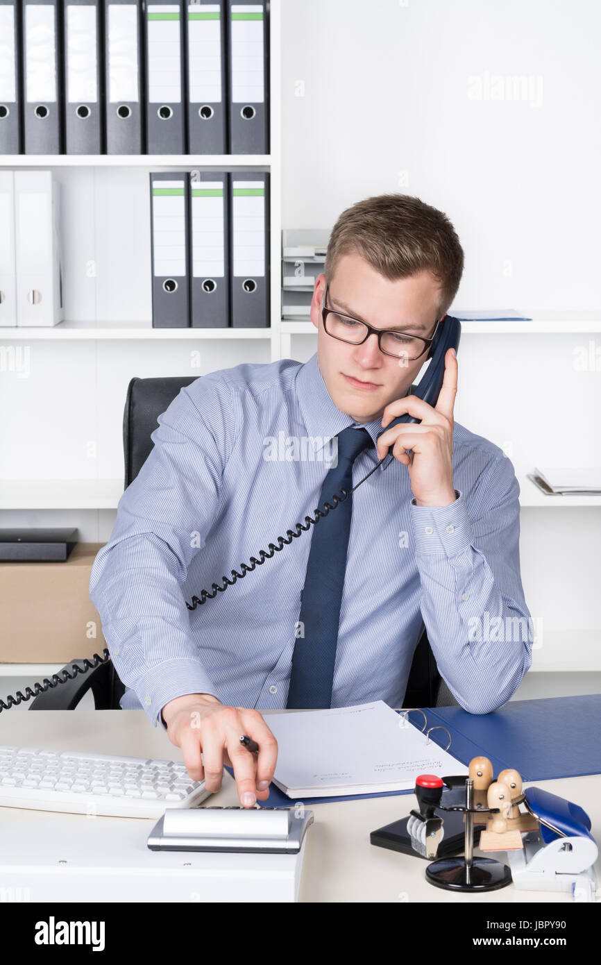Junger Geschäftsmann mit Brille sitzt am Schreibtisch im Büro und telefoniert und tippt auf einem Tischrechner. Im Hintergrund befindet sich ein Regal. Der Mann schaut zum Rechner. Stock Photo