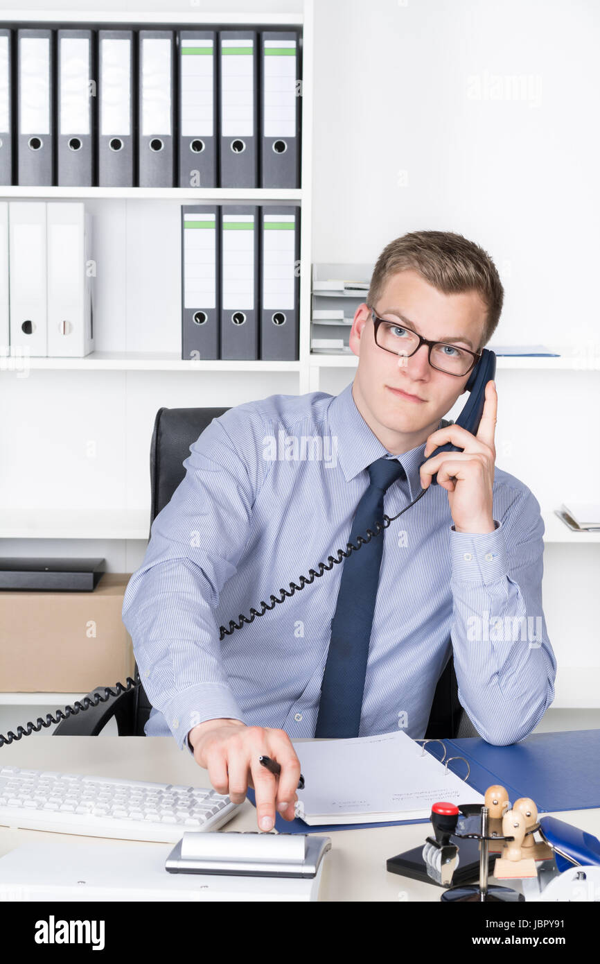 Junger Geschäftsmann mit Brille sitzt am Schreibtisch im Büro und telefoniert und tippt auf einem Tischrechner. Im Hintergrund befindet sich ein Regal. Der Mann schaut zur Kamera. Stock Photo