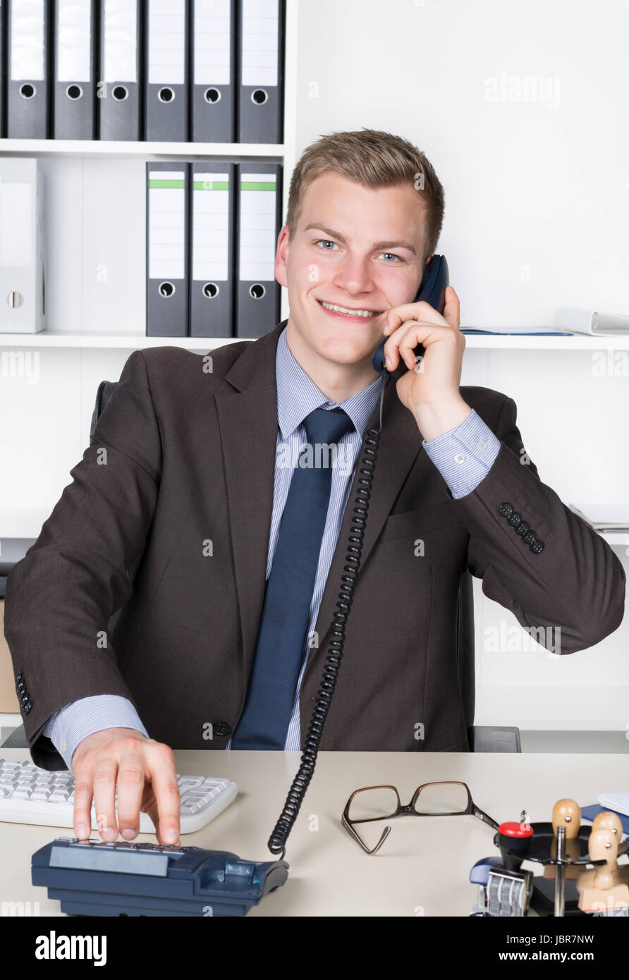 Junger Geschäftsmann sitzt am Schreibtisch im Büro und wählt eine Nummer am Telefon. Im Hintergrund befindet sich ein Regal. Der Mann schaut zur Kamera und lächelt. Stock Photo